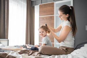 Jeune mère peignes sa peu fille cheveux tandis que séance dans lit dans le Matin photo