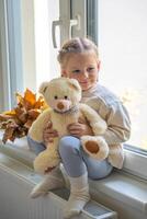 peu fille dans chaud blanc chandail séance par le fenêtre avec peu nounours ours dans l'automne temps photo