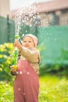 content en riant enfant fille 2-3 année vieux portant imperméable vêtements avoir une amusement avec l'eau pistolet sur Accueil arrière-cour photo