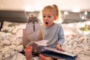 peu fille en train de lire pop en haut livre dans Accueil lit dans Noël environnement avec lumières Contexte. haute qualité photo