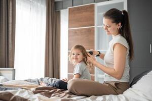 Jeune mère peignes sa peu fille cheveux tandis que séance dans lit dans le Matin photo