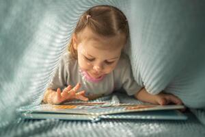 peu fille profiter de la revue une livre en dessous de bleu tricoté plaid dans ensoleillé Matin photo