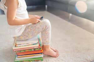 peu fille est assis sur une empiler de enfants Conte de fée livres et montres les dessins animés sur téléphone intelligent à Soleil lumières photo