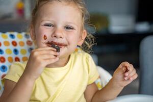 peu fille avec blond cheveux en mangeant fait maison Chocolat avec sale bouche et mains dans Accueil cuisine photo