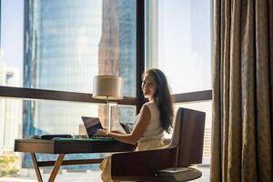 élégant femme d'affaires pigiste travail avec ordinateur portable, elle est séance sur le chaise, panoramique vue dans fenêtre avec ville Contexte. faible clé photo. haute qualité photo