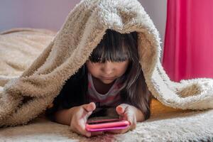 une enfant en utilisant une téléphone intelligent mensonge dans lit, en jouant Jeux, en train de regarder en ligne vidéos, défilement le filtrer. enfants écran dépendance. enfants chambre. photo