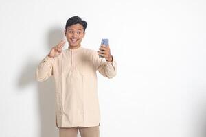 portrait de Jeune excité asiatique musulman homme dans koko chemise en portant mobile téléphone, prise image de lui-même ou selfie, en disant salut et agitant le sien main. social médias concept. isolé sur blanc Contexte photo