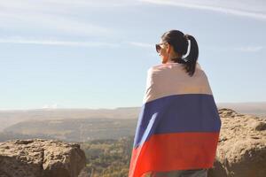 une fille touristique avec une russe drapeau sur sa épaules des stands sur le Haut de une Montagne et jouit le magnifique vues de le Caucase montagnes. photo