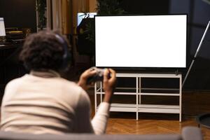 homme en jouant jeux vidéos sur isolé écran la télé, relaxant après longue journée à travail. compétitif joueur ayant amusement sur maquette afficher jeu console dans élégant appartement vivant pièce photo