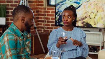 africain américain homme à social rassemblement parlant avec le sien petite amie sur canapé tandis que profiter verre de du vin. mari et épouse invités à appartement fête ayant agréable conversation dans brique mur Accueil photo