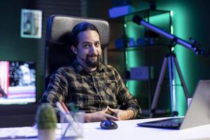 Masculin pigiste à la recherche à caméra, souriant, assise confortablement à une bureau avec le sien portable proche par. un image de une content, barbu homme en utilisant moderne technologie, avec une portable repos sur une tableau. photo