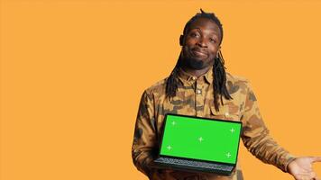 Jeune gars montrant PC avec écran vert disposition sur caméra, montrer du doigt à isolé chromakey afficher. africain américain homme en présentant portable avec Vide fond maquette écran dans studio. photo