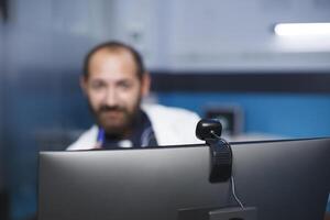 moderne ordinateur étant utilisé par médecin à une hôpital pour une vidéo appel. caucasien homme est assise dans le clinique bureau, participant dans une virtuel conférence à propos avancement de soins de santé traitements. photo