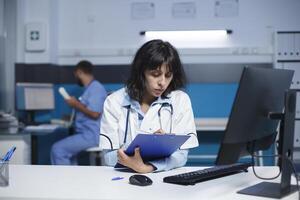 femelle médecin en train de préparer pour médical consultations en utilisant une presse-papiers et ordinateur. image spectacles caucasien femme portant une laboratoire manteau et l'écriture vers le bas Remarques dans une clinique bureau. photo