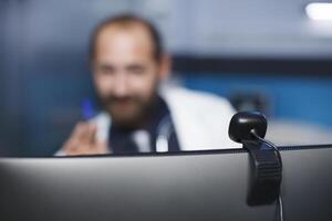 fermer sur ordinateur étant utilisé pour une vidéo bavarder par une médecin dans une hôpital. caucasien homme est assis dans le clinique bureau, engagé dans une virtuel conférence. floue arrière-plan, premier plan se concentrer. photo