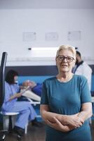 vieux femme dans portrait attend à travail en dehors pour physique traitement avec bras franchi. Sénior patient avec musclé blessure obtient prêt pour jambe physiothérapie sur Stationnaire bicyclette tandis que elle regards à caméra. photo