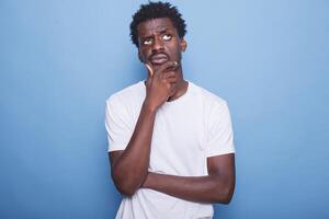 portrait de africain américain gars ayant Profond pensées en portant paume de main à le sien visage à la recherche perdu. noir gentilhomme dans blanc T-shirt rêverie en pensant à propos décontractée journée problèmes. photo