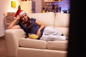 adulte la personne mensonge sur canapé en train de regarder Noël divertissement film sur télévision profiter hiver saison dans Noël décoré cuisine. femme avec rouge Père Noël chapeau célébrer période de Noël photo