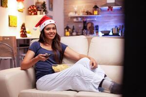 femme souriant tandis que en train de regarder Noël comédie film sur télévision repos sur canapé dans Noël décoré cuisine. fille profiter hiver saison pendant période de Noël célébrer Noël vacances photo