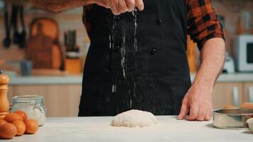 Sénior boulangerie homme tamisage blé farine sur pâte pour pétrissage. retraité personnes âgées chef avec bonte et uniforme saupoudrage, tamisage diffusion rew Ingrédients avec main cuisson fait maison Pizza et pain. photo