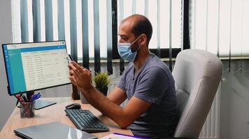 homme séance dans Bureau pièce sur bureau dactylographie sur téléphone portant protection masque. pigiste travail dans Nouveau Ordinaire espace de travail bavardage parlant l'écriture en utilisant mobile téléphone avec l'Internet La technologie photo