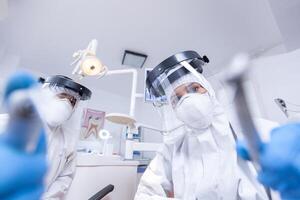 patient pov de dentaire équipe penché plus de à travail dans les dents cavité habillé dans combinaison. stomatologie équipe portant sécurité équipement contre coronavirus traiter patient. photo