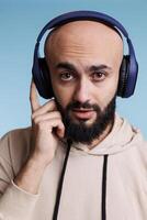 Jeune arabe homme écoute à la musique dans sans fil écouteurs portrait. chauve barbu la personne profiter airs playlist dans écouteurs tandis que à la recherche à caméra avec neutre expression photo