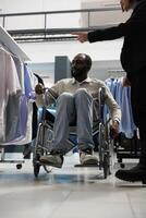 africain américain homme dans fauteuil roulant montrer du doigt à chemise et demander consultant pour Conseil tandis que achats dans Vêtements magasin. boutique client avec invalidité et employé choisir vêtements photo