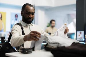 africain américain homme en portant chemise, examiner style et en tissu qualité dans Vêtements magasin. client à la recherche à formel vêtements sur cintre tandis que achats dans centre commercial vêtements pour hommes département photo