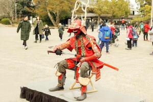 Osaka, Japon, 2023- fermer Humain agissant dans ancien Japonais rouge guerrier armure à Osaka Château parc avec foule de gens et touristique à la recherche autour. photo