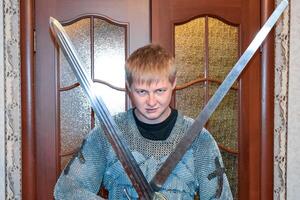 une homme habillé en haut dans une chevalier haubert à Accueil dans le sien chambre. chevalier épée dans le mains de une homme. photo