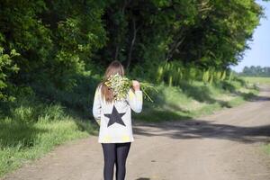 une fille en marchant le long de une forêt route avec une bouquet de camomille photo