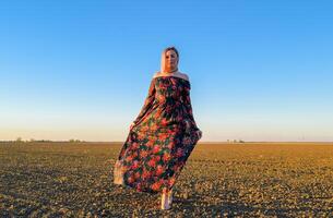 femme dans une labouré champ dans une rouge noir robe sur une le coucher du soleil Contexte. photo