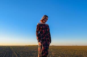 femme dans une labouré champ dans une rouge noir robe sur une le coucher du soleil Contexte. photo