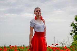 blond Jeune femme dans rouge jupe et blanc chemise, rouge des boucles d'oreilles est dans le milieu de une coquelicot champ. photo