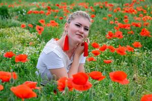 blond Jeune femme dans rouge jupe et blanc chemise, rouge des boucles d'oreilles est dans le milieu de une coquelicot champ. photo