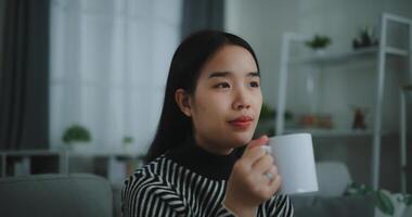 portrait de relaxant Jeune femme en portant tasse prendre plaisir odeur café ou thé et en buvant avec bonheur dans Matin tandis que asseoir sur canapé dans vivant chambre, gratuite le temps, prends Pause dans à la maison, souriant photo