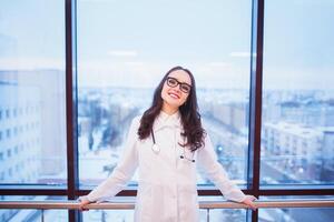 joyeux Jeune infirmière. souriant Jeune fille dans blanc uniforme en portant numérique tablette et à la recherche à caméra, clinique fenêtre Contexte. photo