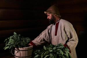 homme dans traditionnel robe prépare une baignoire balais dans une traditionnel vapeur sauna banya photo