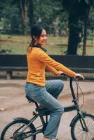 content Jeune asiatique femme tandis que équitation une vélo dans une ville parc. elle sourit en utilisant le vélo de transport. écologiquement amical concept. photo