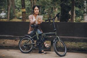 content Jeune asiatique femme tandis que équitation une vélo dans une ville parc. elle sourit en utilisant le vélo de transport. écologiquement amical concept. photo