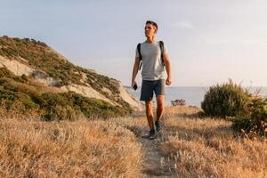 Beau homme avec sac à dos en marchant sur le océan littoral avec montagnes et soir chaud lumière du soleil. photo