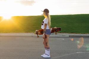 portrait de une branché Jeune fille souriant avec une longboard à le coucher du soleil. photo