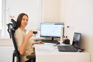 portrait de une éloigné ouvrier. une magnifique souriant brunette fille est assis à une table dans de face de une ordinateur et travaux avec une mise en page. sur de soi Jeune Dame pigiste travail de maison. photo
