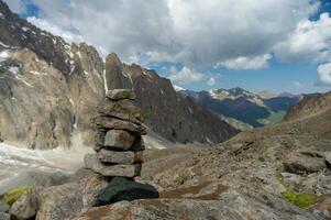 empilés Roche cairn des stands en bonne place contre une toile de fond de imposant saupoudré de neige pics, en dessous de une vaste bleu ciel avec épars des nuages, surplombant une vallée avec conseils de vert feuillage. photo