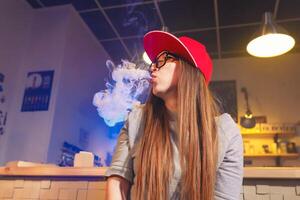Jeune jolie femme dans rouge casquette fumée un électronique cigarette à le vape magasin photo