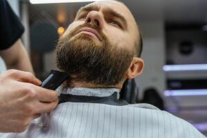 coiffeur garniture barbu homme avec rasage machine dans salon de coiffure. coiffure processus. fermer de une coiffeur Coupe le barbe de une barbu Masculin. photo