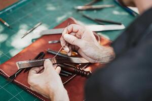 fermer main travail processus cuir artisanat dans le cuir atelier. homme en portant artisanat outil et fonctionnement. il est couture à faire une portefeuille. tanneur dans vieux tannerie. photo