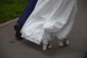 le ourlet de une dentelle blanc mariage robe et le du marié jambes. le jeunes mariés marcher le long de le asphalte chemin dans le parc. photo