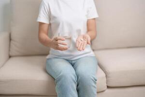 femme main en portant médicament anti douleur pilule et l'eau verre sur le canapé à maison, prise pour maux de tête, estomac mal, la diarrhée douleur de nourriture empoisonnement, l'endométriose, hystérectomie et menstruel photo